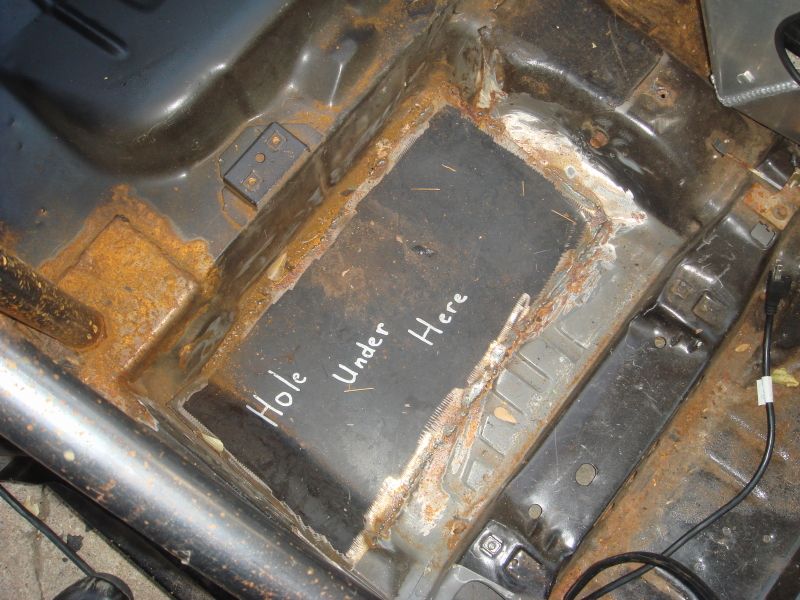 Rusted car floor interior with handwritten text 'Hole Under Here'