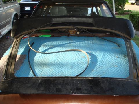 1989 Ford Escort - The rear window frame of a car is shown removed and placed on a blue quilted mat, with other vehicles in the background.