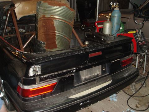 1989 Ford Escort - Modified 1989 Ford Escort rear view shows a roll cage and a large rusty barrel in the trunk, with welding equipment to the side.