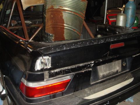 1989 Ford Escort - The rear of a black car, modified with a roll cage and carrying a rusty barrel, is shown.
