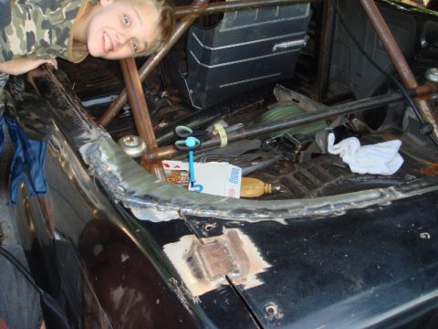1989 Ford Escort - A child smiles while looking over the fender of a car being worked on, revealing its interior roll cage and some tools.