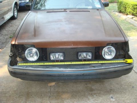 1989 Ford Escort - A tape measure is placed across the front of a brown car, obscuring the headlights and grille.