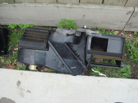 1989 Ford Escort - A disassembled HVAC blower motor housing rests outdoors near a wooden fence.