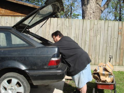 1989 Ford Escort - A person is seen loading items into the open trunk of a dark colored hatchback car.