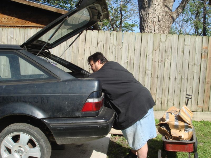 Man loading items into the trunk of a dark hatchback. Text on the car reads "MONSCORT"