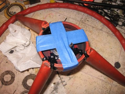 1966 Ford F-100 - A red three-spoke steering wheel lying face-up on a workbench during repair, with blue painter's tape crossed over the center hub and black…