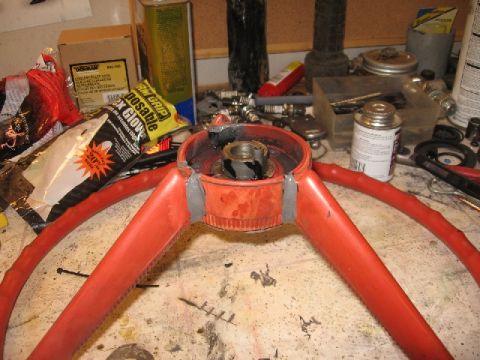 1966 Ford F-100 - A red three-spoke steering wheel laid face-down on a cluttered workbench, showing the splined hub and center boss with masking tape wrapped…