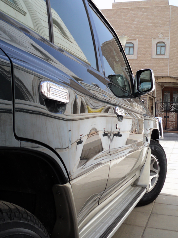 Close-up of dark SUV's reflective side panels, mirror, and wheel parked by a beige building.