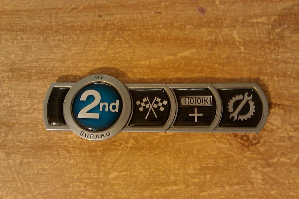 Chrome "My 2nd Subaru" badge with checkered flag, 100K+ and wrench icons on wood