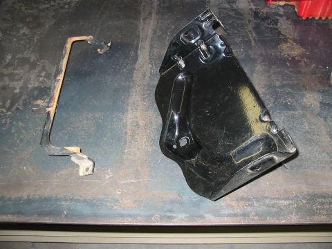 1993 Ford Taurus - Two removed black-painted stamped steel brackets laid out on a scarred workbench: a long slender strap-style mounting bracket at left and a…