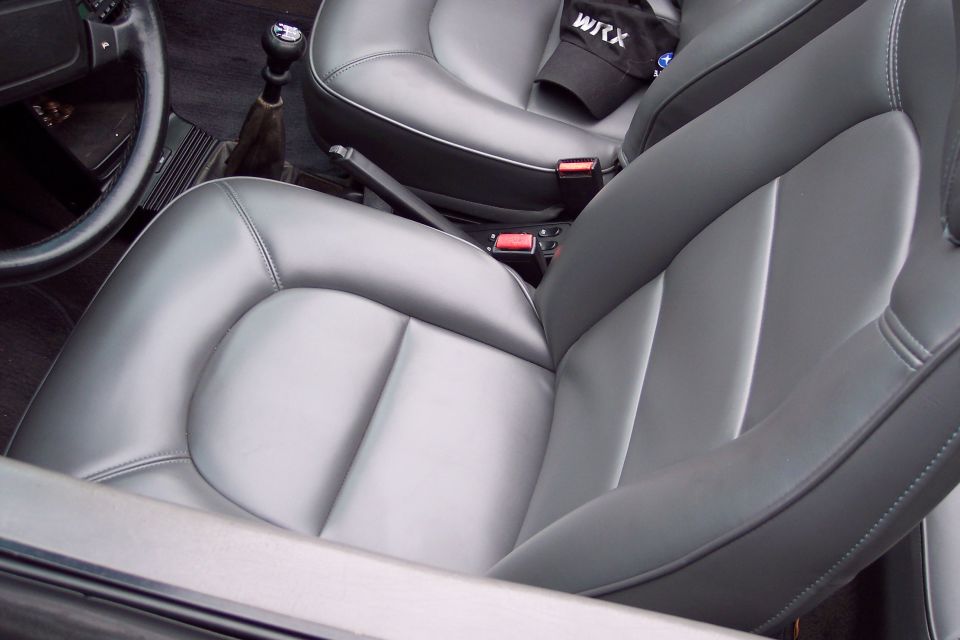Interior view of car seats, gear shifter, and steering wheel. A black cap with 'WRX' is on the passenger seat