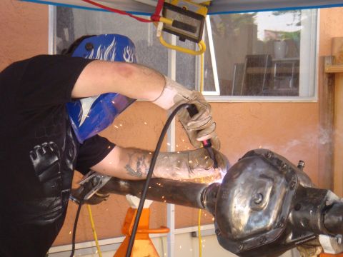 1992 Ford Bronco - A person in a welding mask and gloves works on a vehicle axle, with sparks emanating from the welding torch.