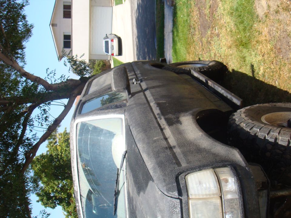 Black Ford Bronco parked in a driveway, tree branches overhead. Another white SUV is parked in the garage behind it