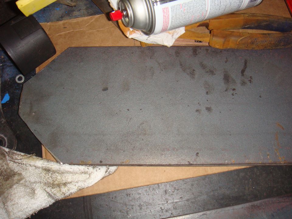 Flat metal part with angled edges on a workbench, next to a spray can and rags