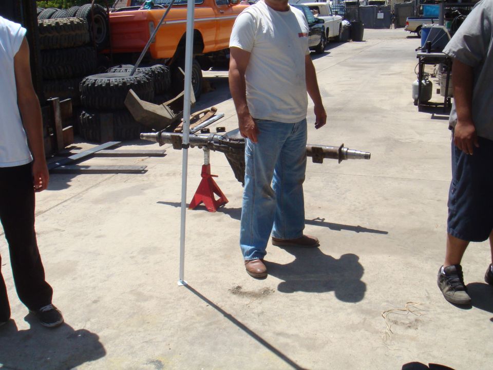 Vehicle axle assembly on red jack stands, with an orange truck and stacked tires in the background