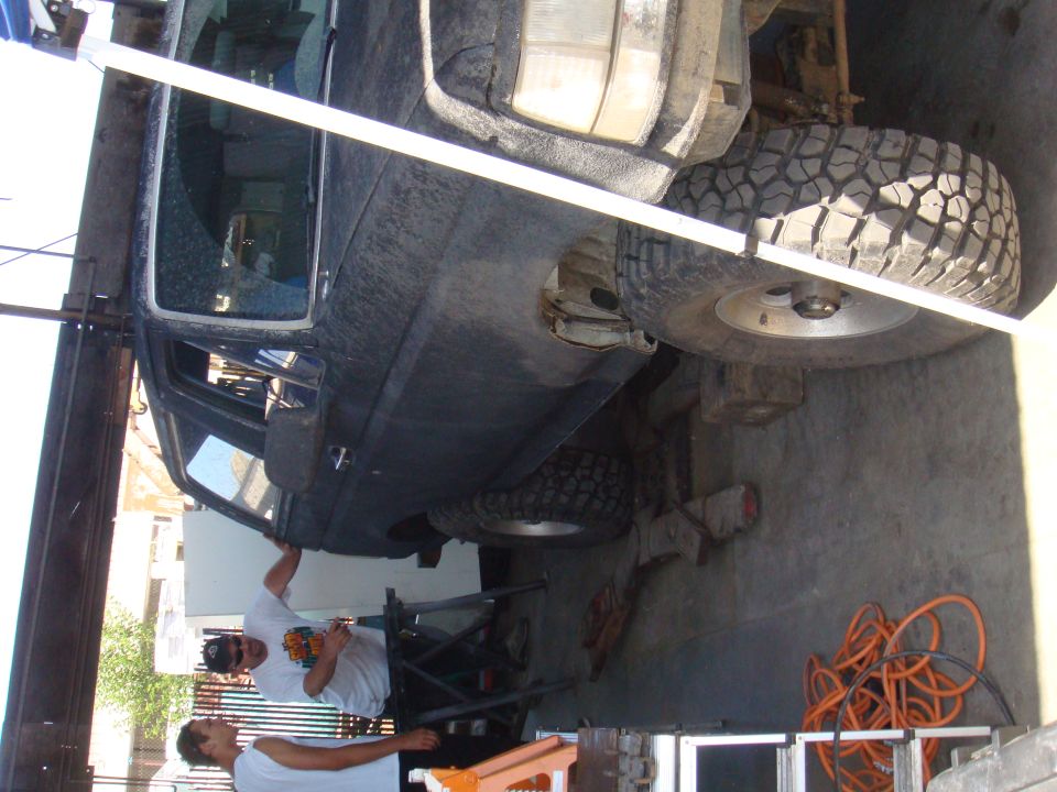 Side view of dark SUV on lift with men working underneath. No year visible