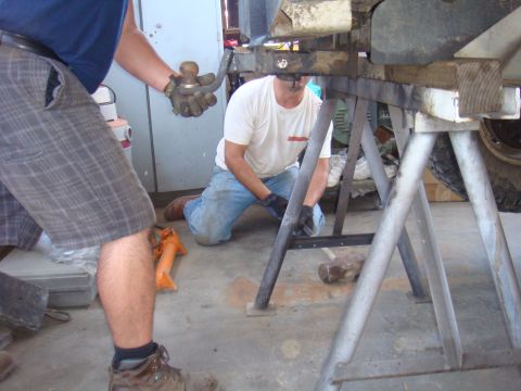 1992 Ford Bronco - Two individuals are seen working on a vehicle's frame, which is elevated on stands. A tow hitch component is held by one person.