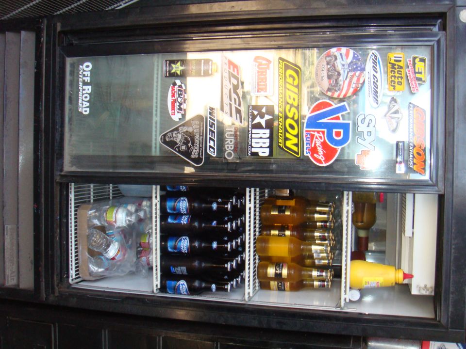 Refrigerator door covered in various automotive stickers; interior stocked with beer bottles and mustard