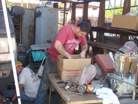 1992 Ford Bronco - Two mechanics are working on a vehicle in a cluttered outdoor workshop. Tools and parts are spread across a table.