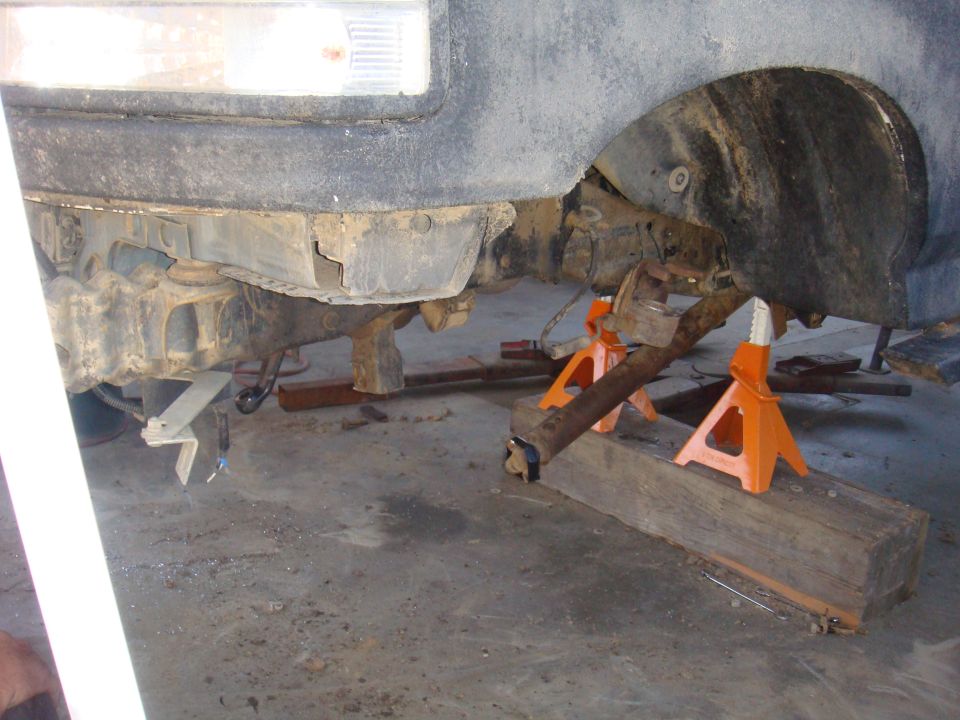 Vehicle's rear axle and suspension components are supported by jack stands and wooden blocks in a garage