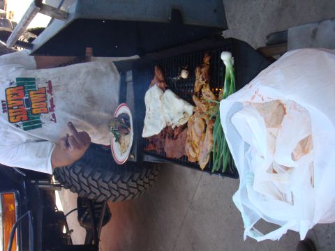 1992 Ford Bronco - A person grills food next to a large tire, with a white plastic bag full of items nearby.