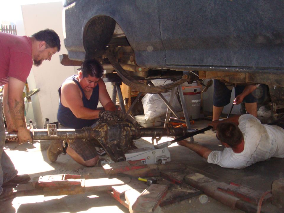 Three men working on a vehicle's rear axle assembly, which is suspended by jacks