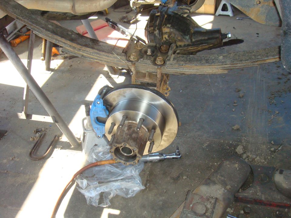 Vehicle rear axle with new brake rotor and blue caliper installed. Leaf springs and U-bolts are visible