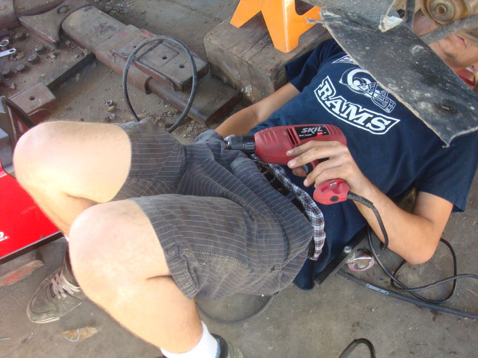 Person wearing a Rams shirt and shorts holds a red Skil drill while working on something under a vehicle