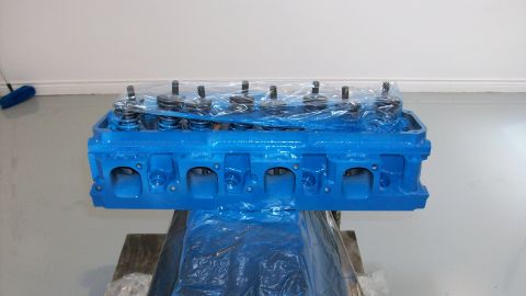 1973 Ford Ranchero - A blue engine cylinder head, wrapped in plastic, sits on a stand in front of a white wall.