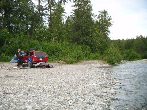 1998 Jeep Cherokee - A red 1998 Jeep Cherokee is parked on a rocky shore beside a river, with camping gear and a tent set up.