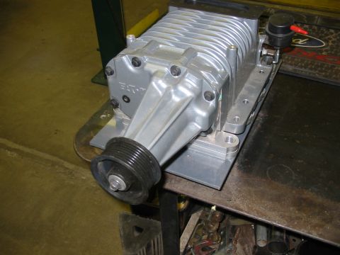 1993 Ford Taurus - A cast-aluminum Roots-type supercharger sits on a steel workbench, mounted to a machined adapter plate with a black six-rib drive pulley on…