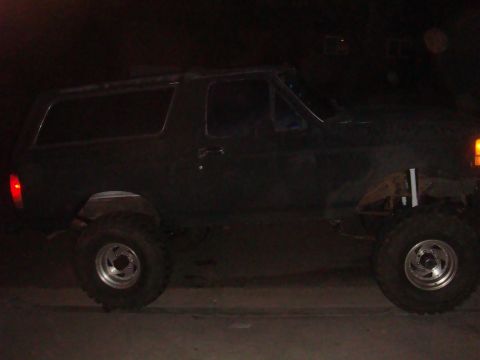 Side view of a dark-colored Ford Bronco, possibly from the early 1980s, with large tires and lifted suspension