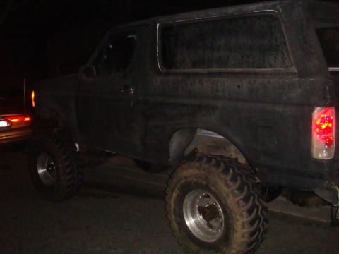 Black Ford Bronco with large tires, seen from the side at night. Another vehicle is partially visible on the l
