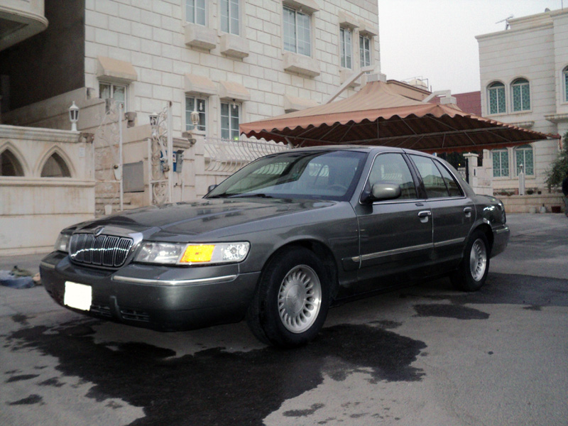 Gray Mercury Grand Marquis sedan, front 3/4 view, parked by stone building
