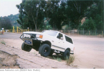 1988 Ford Bronco - White Ford Bronco with custom bumper and off-road tires navigates a dirt incline.