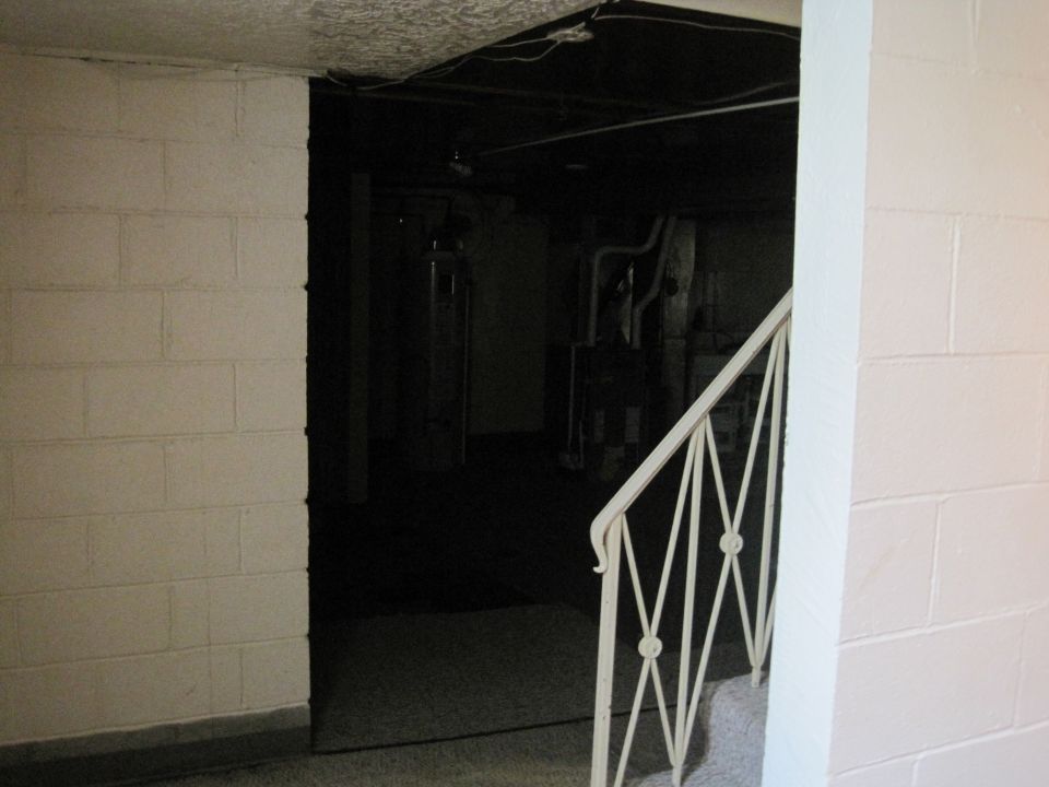 Basement interior with white brick walls, a dark ceiling with exposed wires, and a white metal railing leading down stairs
