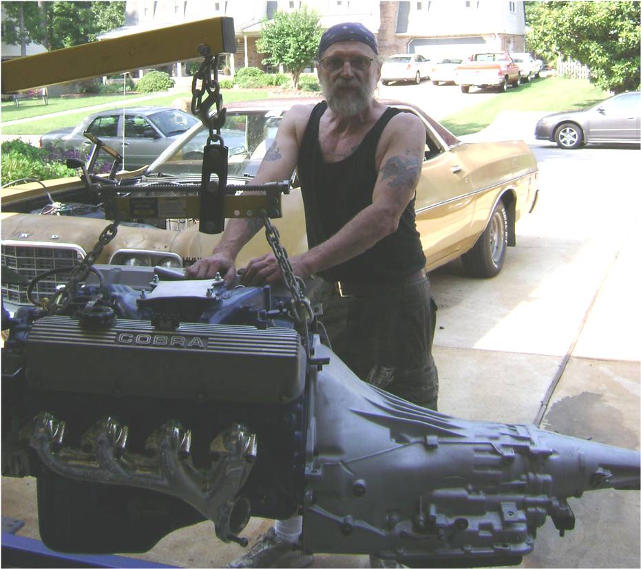 Man beside a Cobra-valve-cover V8 and transmission on a hoist, gold Ranchero behind