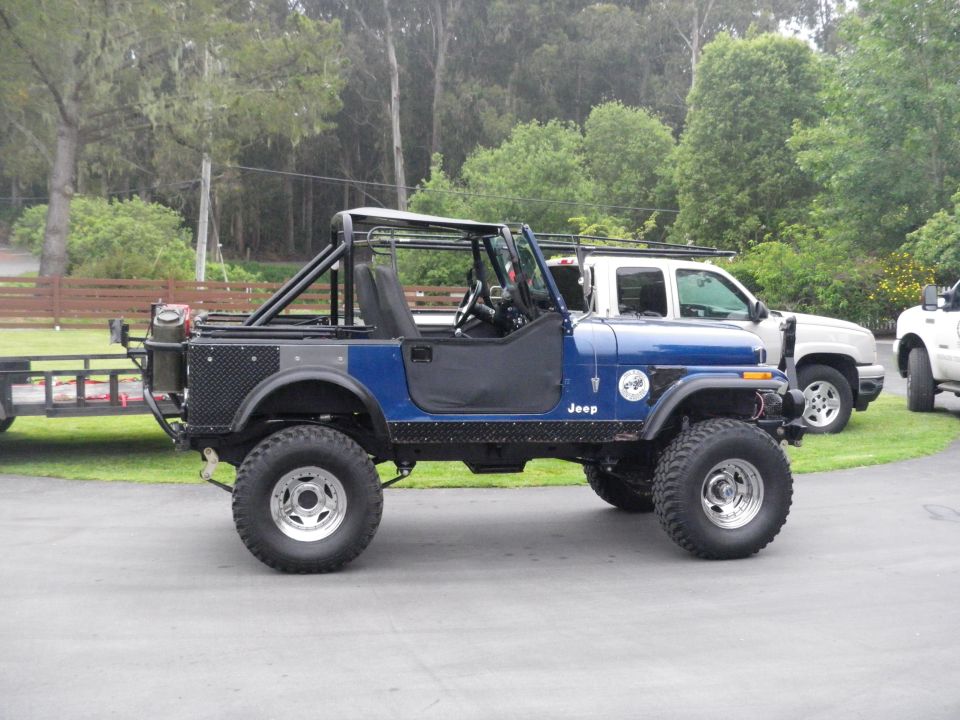 Blue Jeep CJ-7 side view with roll cage and trailer attached, two white trucks in background