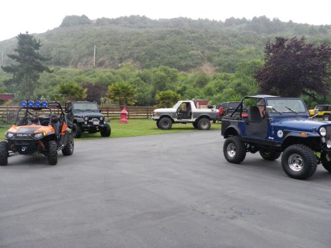 1991 Ford Bronco - A collection of off-road vehicles are gathered in a paved area with a grassy hill in the background.