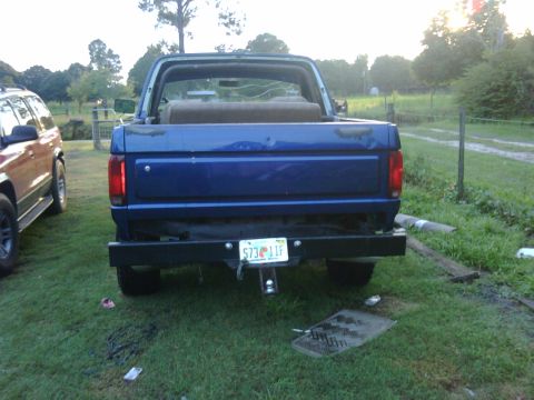 1991 Ford Bronco - The rear of a blue Ford Bronco is visible, parked on grass with a red SUV to its left.