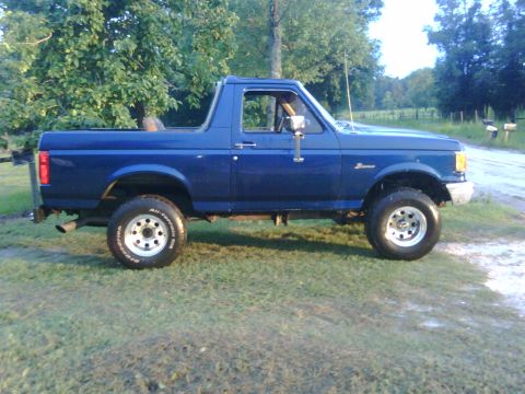 1991 Ford Bronco - This blue 1991 Ford Bronco is shown from the side, parked on a grassy area with trees in the background.