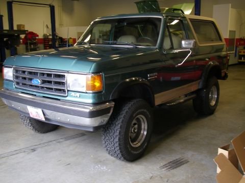 1990 Ford Bronco - Front three-quarter view of a lifted green-over-tan Ford Bronco with a tan hardtop, black fender flares, a 4x4 grille decal, and BFGoodrich…