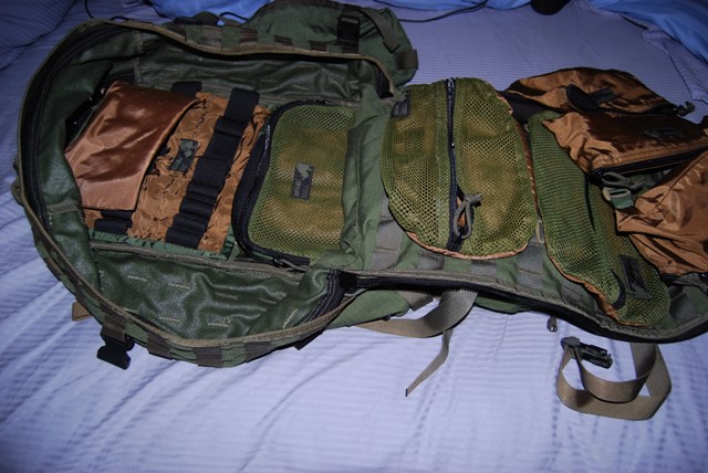 Open green and tan backpack with multiple zippered compartments and straps, laid on a blue striped surface