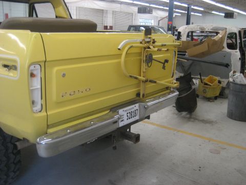 1978 Ford Bronco - The rear of a yellow 1978 Ford Bronco is shown, a spare tire mount and a tow hitch. Another vehicle is visible in the workshop setting.