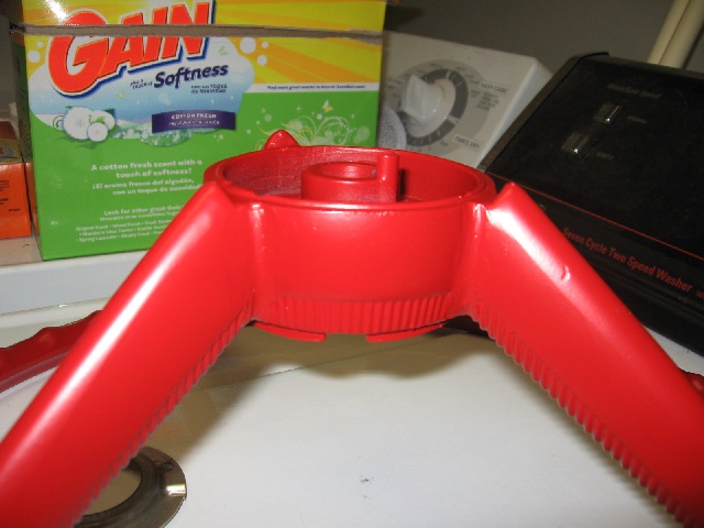 Red plastic steering wheel part with two spokes, resting on a white surface near laundry detergent and a washing machine
