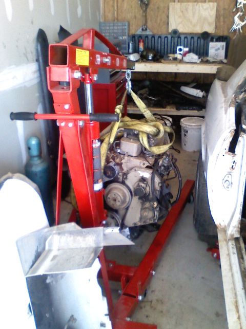 1990 Ford Bronco - An engine is suspended by a red engine hoist in a garage, with a portion of a white vehicle visible to the right.