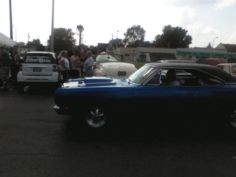 1960 Ford Falcon - A blue coupe with a prominent hood scoop is seen from the side, with other vehicles and people in the background.