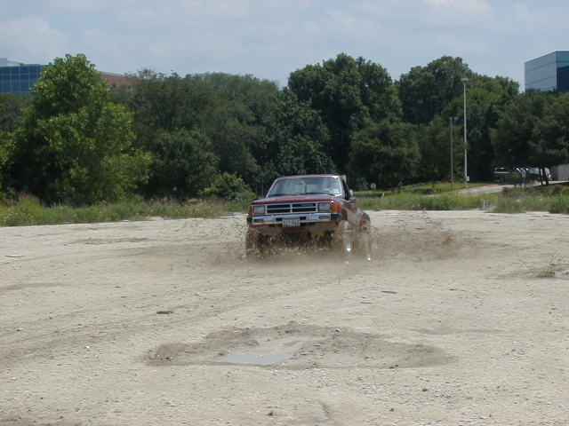 Red pickup truck kicking up mud while driving off-road. The truck appears to be from the 1980s