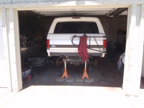 1994 Ford Bronco - Rear view of a white full-size Ford Bronco raised on orange jack stands set on concrete pavers inside an open garage, with both rear wheels…