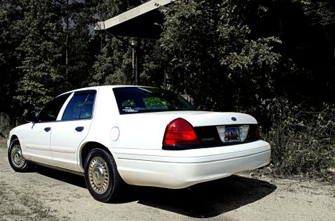 2002 Ford Crown Victoria - Rear view of a white 2002 Ford Crown Victoria parked outdoors with trees and a structure overhead.